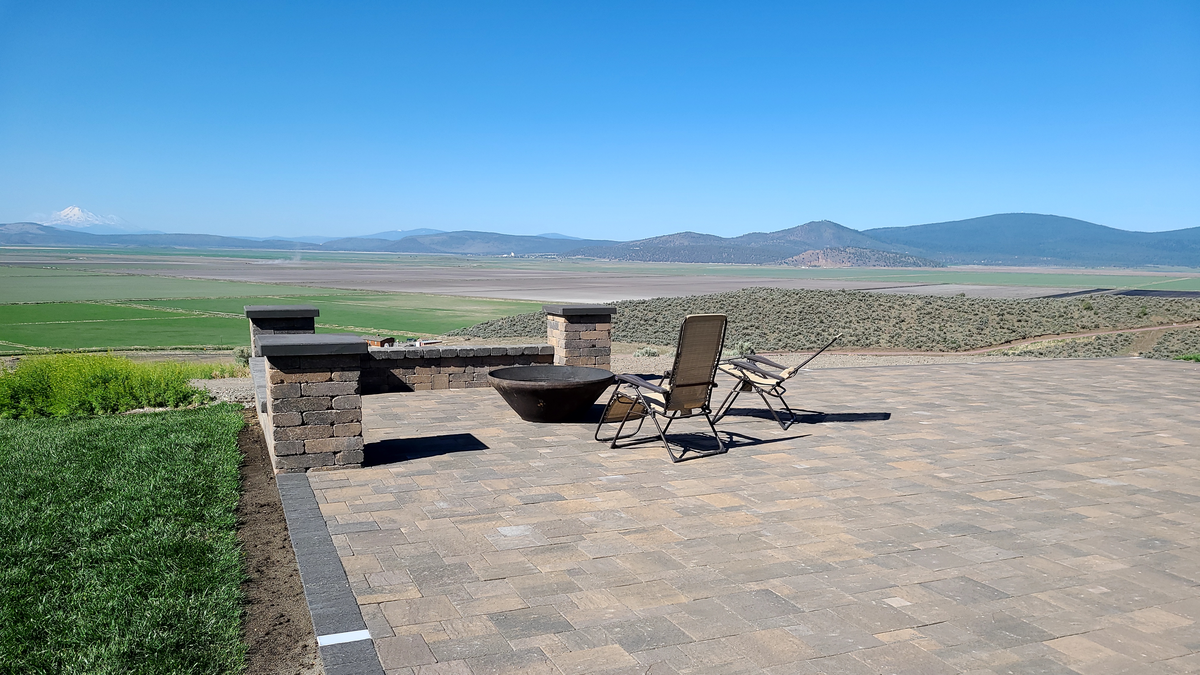 Custom stone patio with paver flooring, fire bowl, and lounge seating with a panoramic view of farmland and distant snow-capped mountains